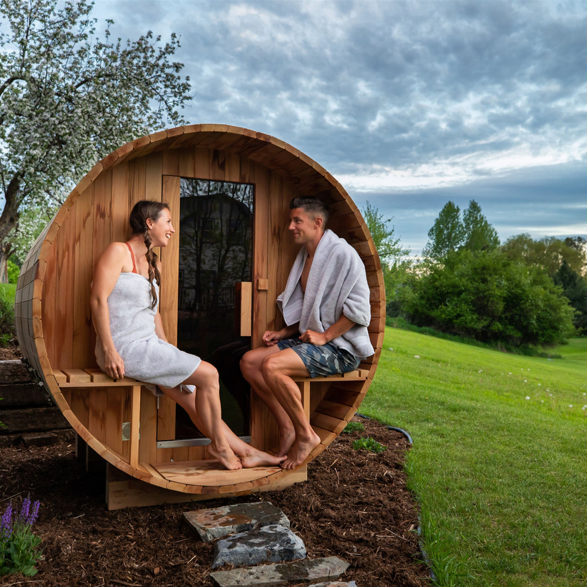 A couple resting on the canopy of Aleko Red Cedar Wet/Dry Outdoor Barrel Sauna with Front Porch Canopy, Panoramic View, Butimen Shingle Roofing and 8 kW KIP Harvia Electric Sauna Heater.