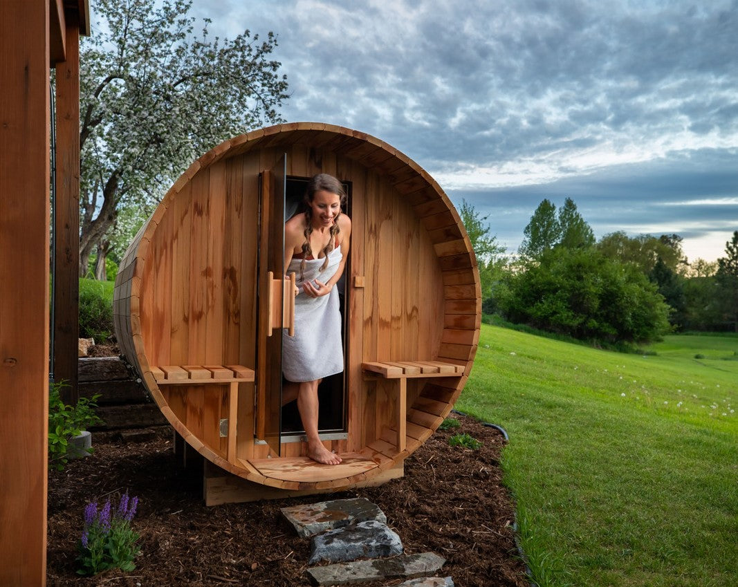 A woman getting out of a Aleko Red Cedar Wet/Dry Outdoor Barrel Sauna with Front Porch Canopy, Panoramic View, Butimen Shingle Roofing and 8 kW KIP Harvia Electric Sauna Heater in a natural setting.