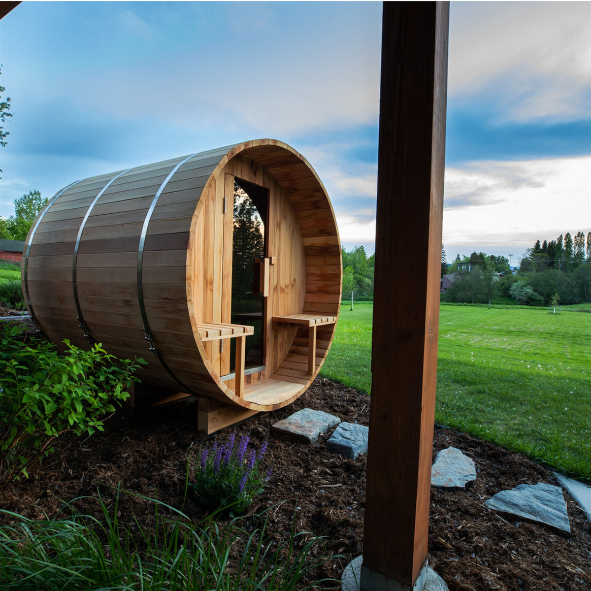 Side view of Aleko Red Cedar Wet/Dry Outdoor Barrel Sauna with Front Porch Canopy, Panoramic View, Butimen Shingle Roofing and 8 kW KIP Harvia Electric Sauna Heater in a natural setting.