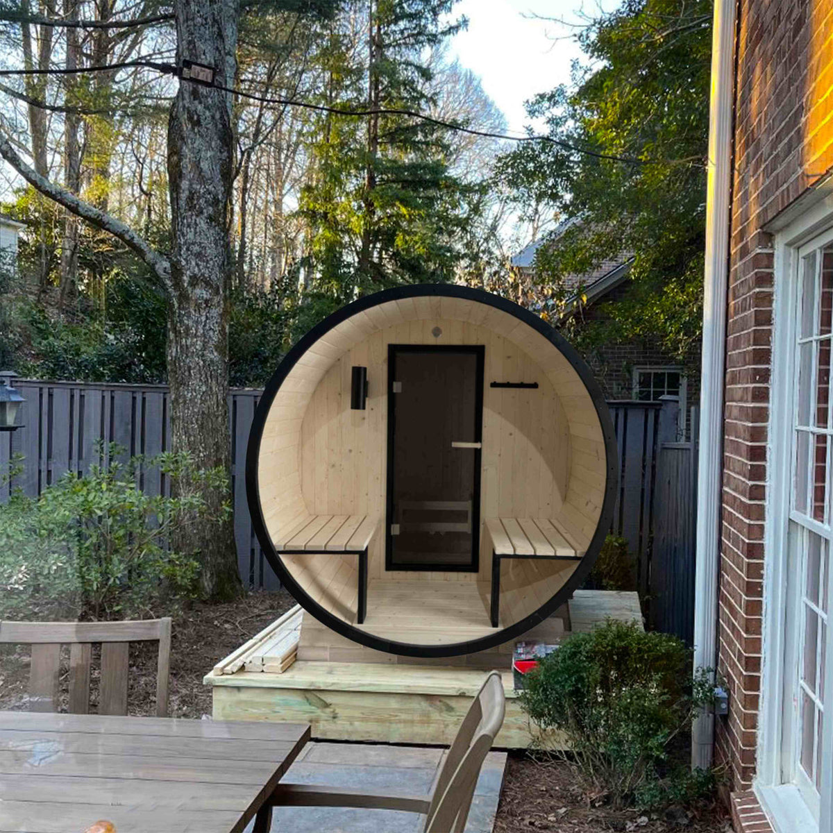 Aleko White Finland Pine 3/5 Person Traditional Outdoor Barrel Sauna with Black Accents, Front Porch Canopy and 4.5 kW Harvia KIP Electric Sauna Heater in a garden.