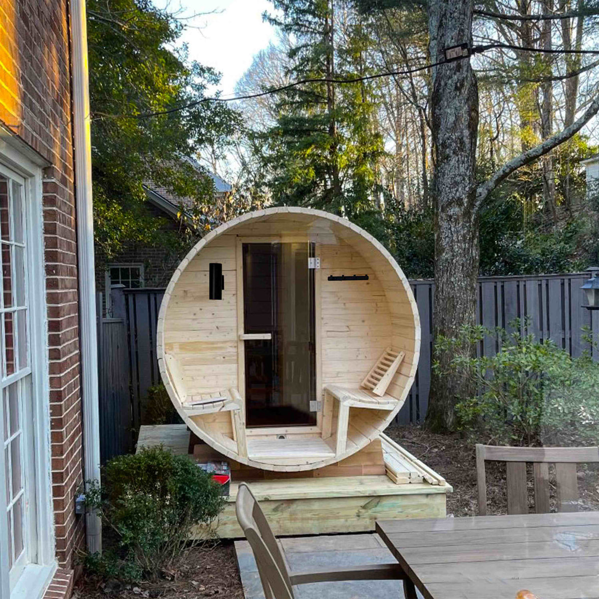 Front view of Aleko White Finland Pine Wet/Dry 6/8 Person Outdoor Barrel Sauna with Front Porch Canopy and 8 kW Harvia KIP Electric Sauna Heater in a garden.