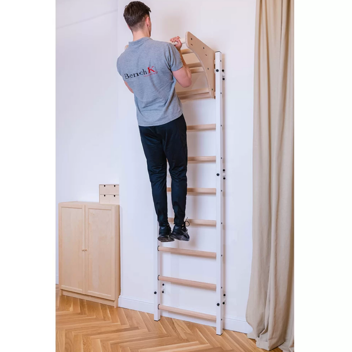 A man exercising with a BenchK 711 Wall Bars and Gymnastic Ladders with wooden pull-up bar white version installed in a bedroom.