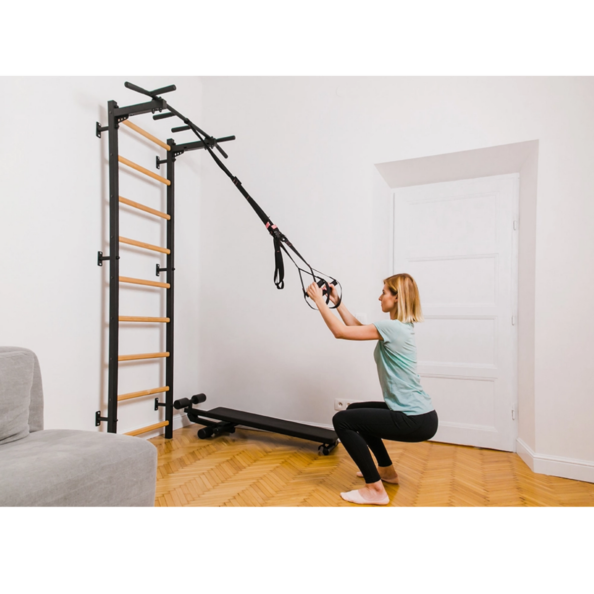 A woman exercising with a TRX attached to BenchK 721 Wall Bars + A076/A204 Gymnastic Accessories black version installed in a living room.