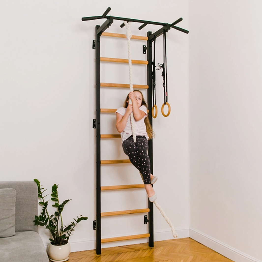 A kid playing with the rope of BenchK 721 Wall Bars + A076/A204 Gymnastic Accessories black version installed in a living room.