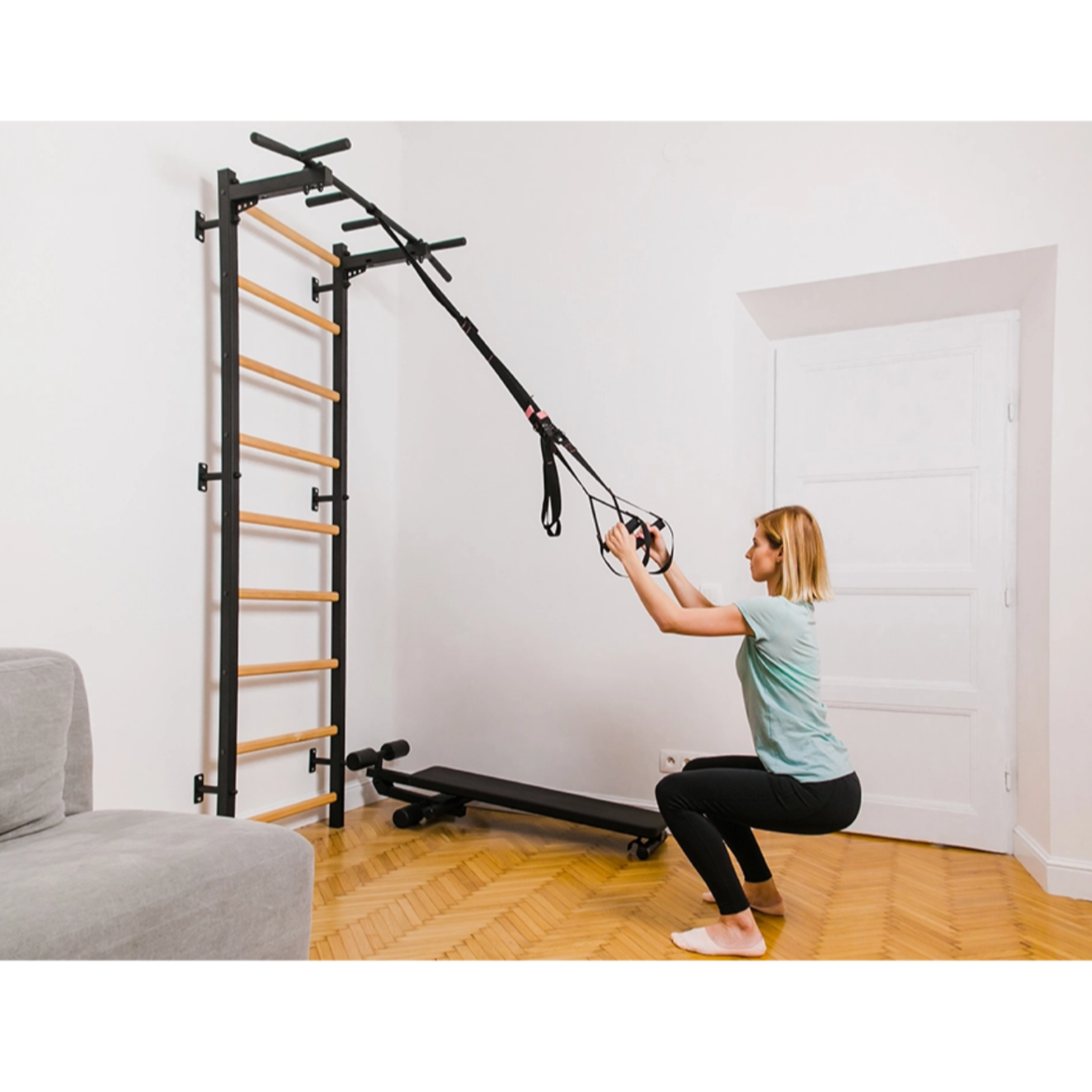 A woman exercising with a TRX attached to BenchK 721 Wall Bars and Gymnastic Ladders black version installed in a living room.