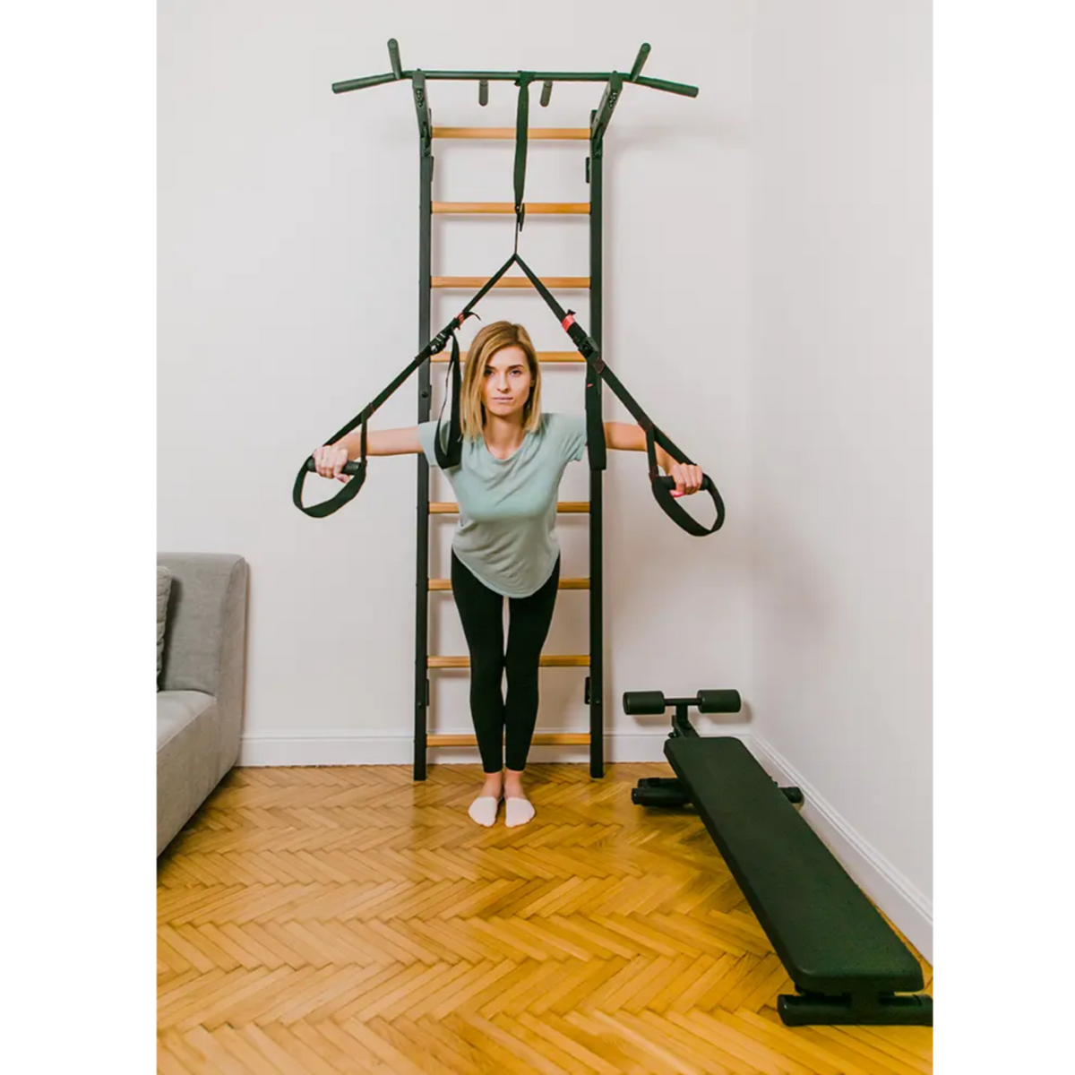 A woman exercising with a TRX attached to BenchK 721 Wall Bars and Gymnastic Ladders black version with pull-up bar installed in a living room.