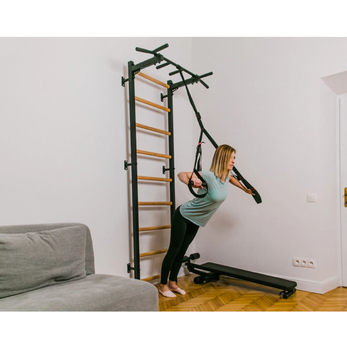 A woman exercising with a TRX attached to BenchK 721 Wall Bars and Gymnastic Ladders black version installed in a living room.