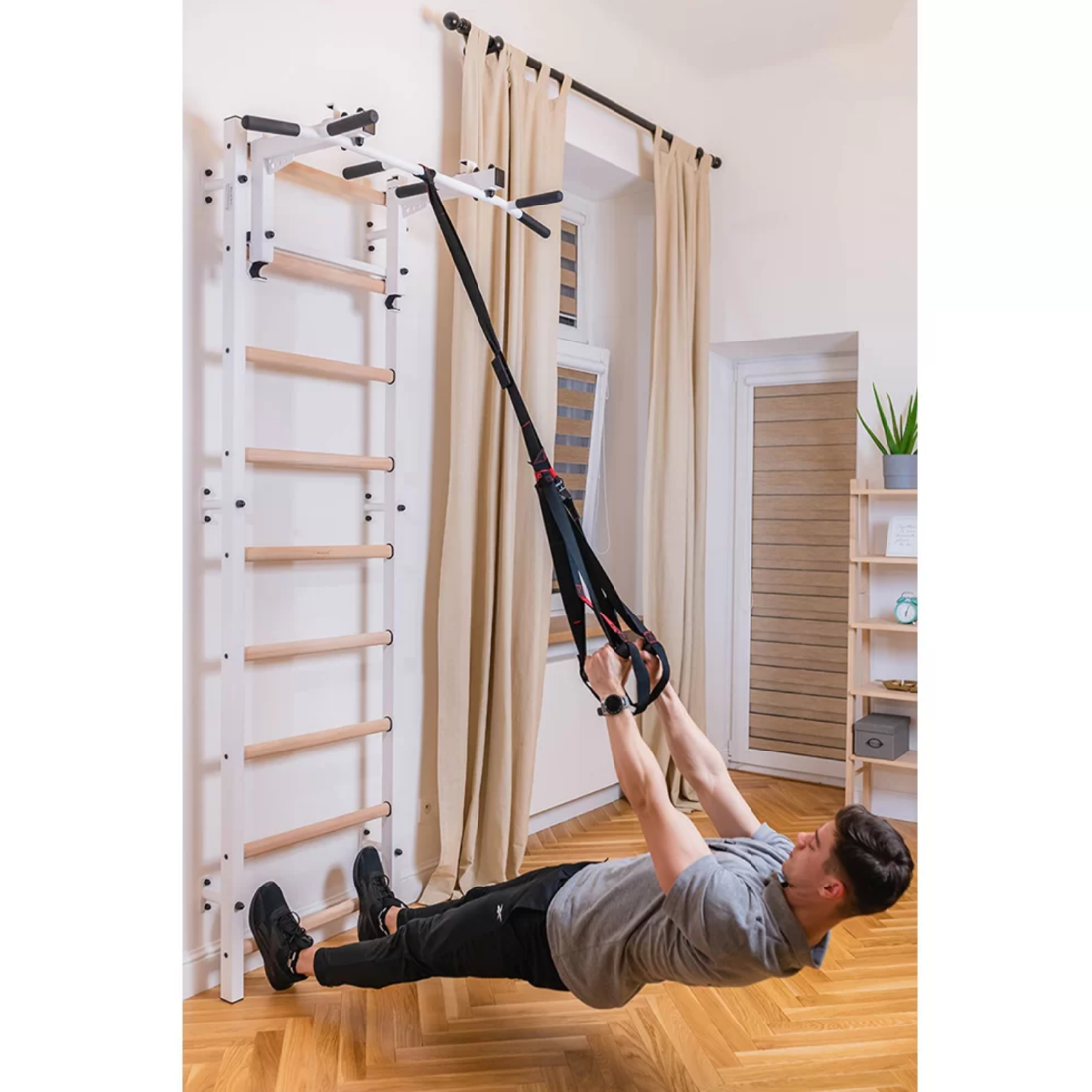 A man using a TRX with a BenchK 731 Wall Bars and Gymnastic Ladders white version installed in a living room.