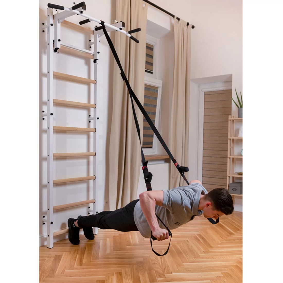 A man exercising with a TRX attached to BenchK 731 Wall Bars and Gymnastic Ladders white version installed in a living room.