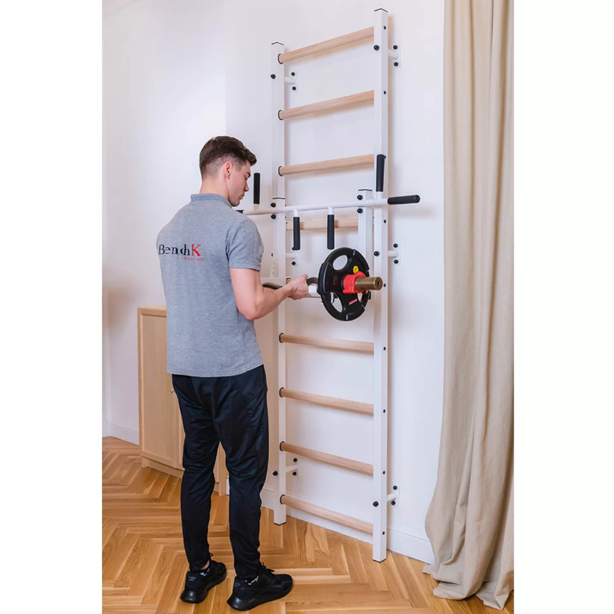 A man taking a barbell from the BenchK 731 Wall Bars and Gymnastic Ladders white version installed in a living room.