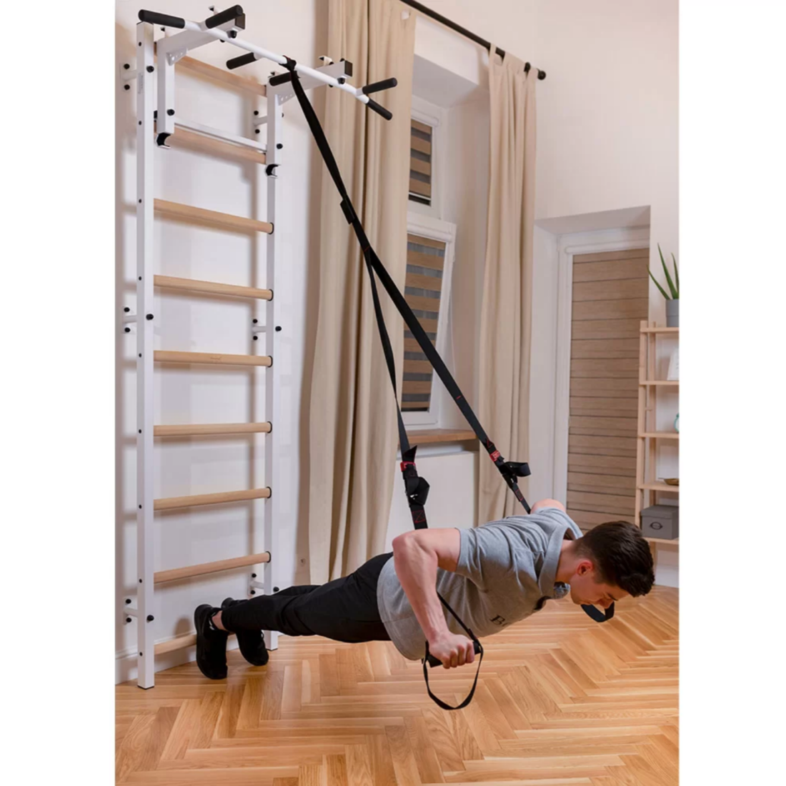 A man exercising with a TRX attached to BenchK 732 Wall Bars and Gymnastic Ladders white version installed in a bedroom.