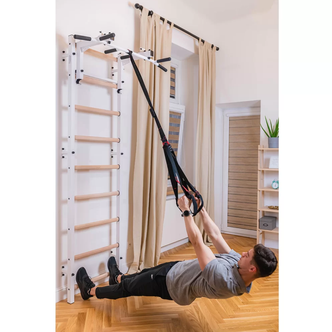 A man exercising with a TRX attached to BenchK PB3 Steel Pull-Up Bar with barbell holder white version installed in a living room.