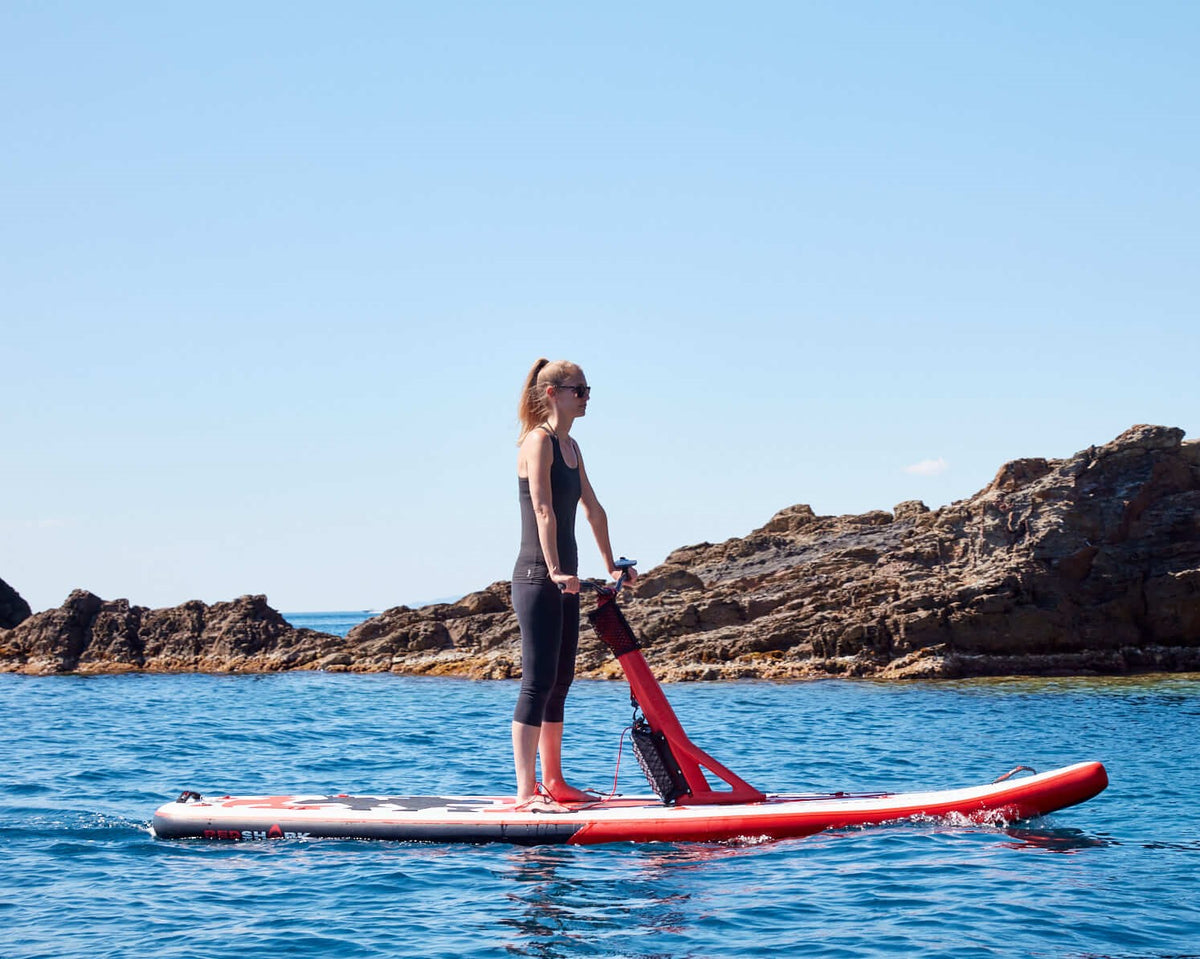 Female rider on a Red Shark Scooter Bike Surf near rocky coastline under clear blue sky.