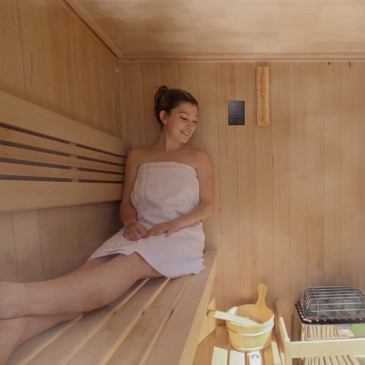 Woman relaxing in wooden sauna with Harvia KIP Electric Sauna Heater 8 kW and WiFi Remote Control Panel on wall.