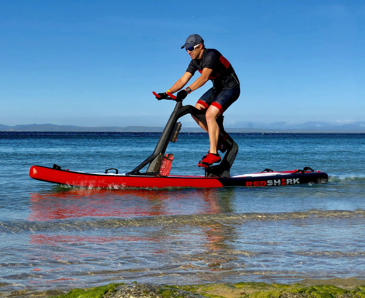 Man pedaling a Red Shark Fitness Bike Surf along the shoreline with clear blue water.