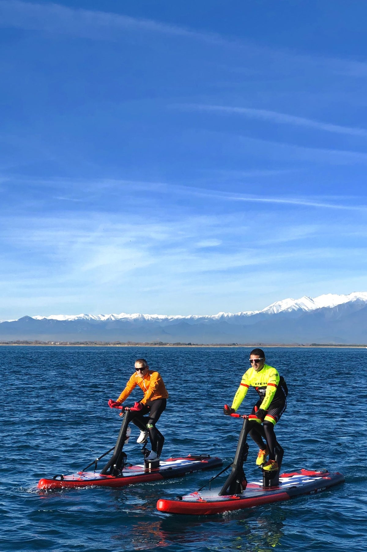 Two riders on Red Shark Fitness Bike Surfs on the sea with a stunning mountain backdrop.