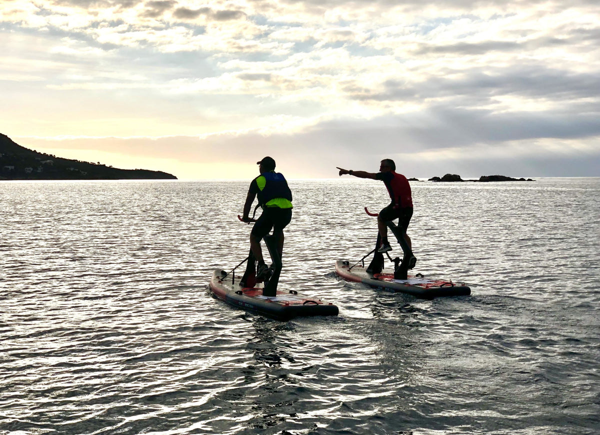 Silhouettes of two Red Shark Fitness Bike Surf riders on the sea at sunset with islands in the distance.