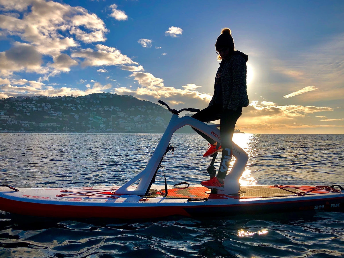 Silhouette of a person on a Red Shark Enjoy Bike Surf at sunset with cityscape in the background.